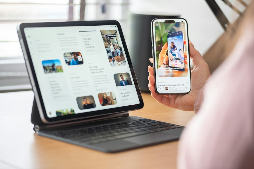 Person holding a smartphone displaying a video thumbnail while a tablet with app cards sits on a keyboarded laptop on a desk.