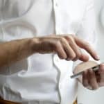 Person in a white shirt taps on a white smartphone held in one hand.