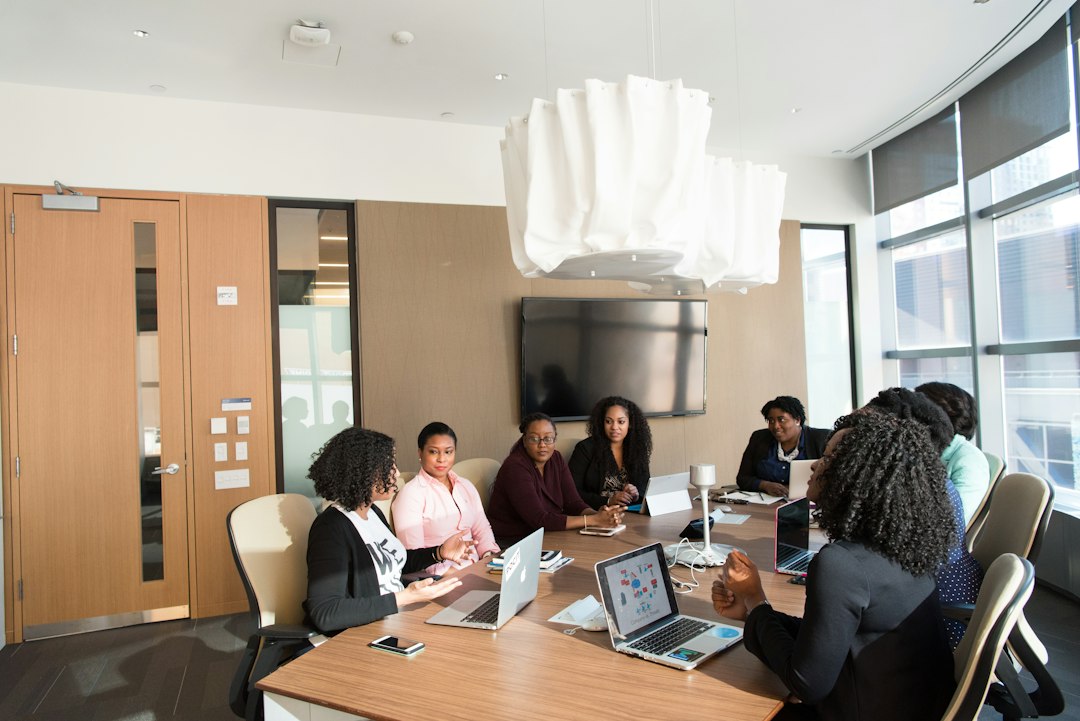 A diverse group of professionals seated around a conference table with laptops, in a bright meeting room with a large window.