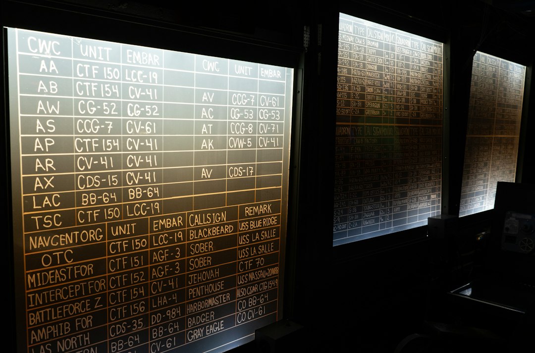 Backlit chalkboard wall filled with rows of vessel codes and assignment columns (CWC, UNIT, EMBAR) in a dim room with multiple boards along the wall.