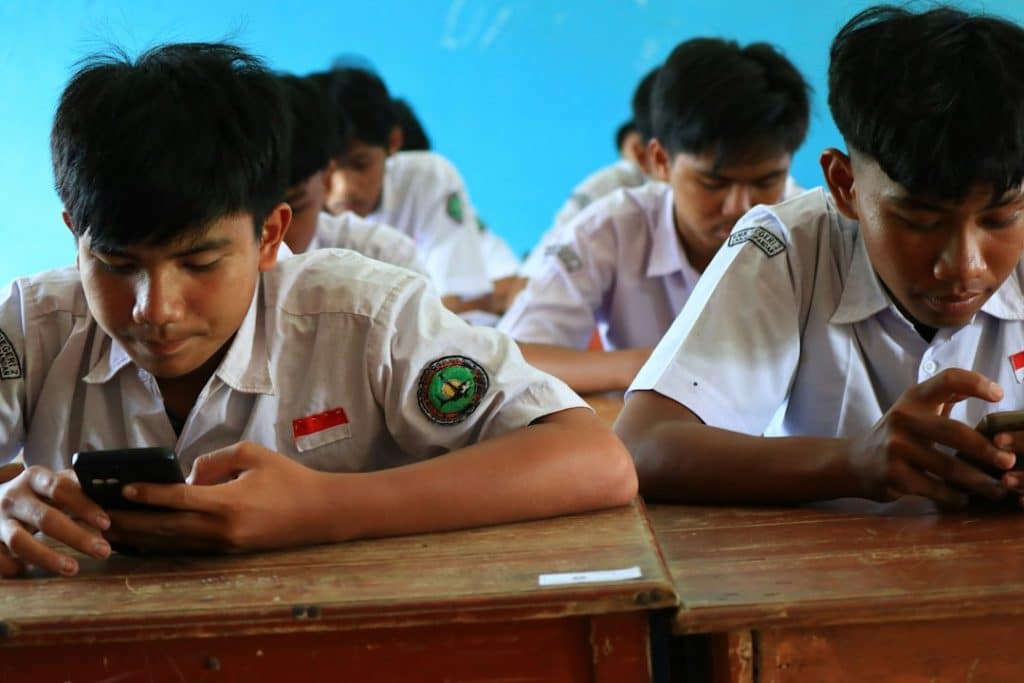 Two male students in white uniforms sit at a wooden desk in a classroom with a blue wall, each looking at a smartphone.