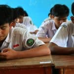 Two male students in white uniforms sit at a wooden desk in a classroom with a blue wall, each looking at a smartphone.