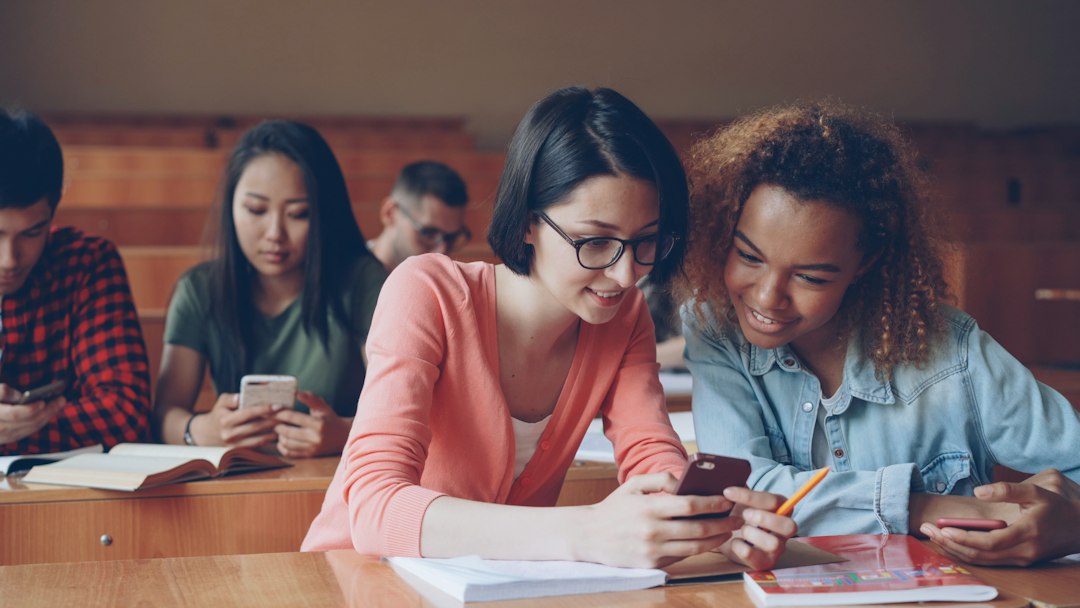 Two students share a smartphone at a classroom desk while others study with phones in the background.