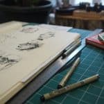Open sketchbook on a green cutting mat with pen, ruler, and markers scattered nearby in an artist's workspace.