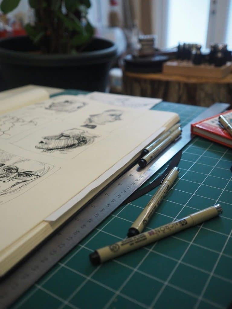 Open sketchbook on a green cutting mat with pen, ruler, and markers scattered nearby in an artist's workspace.