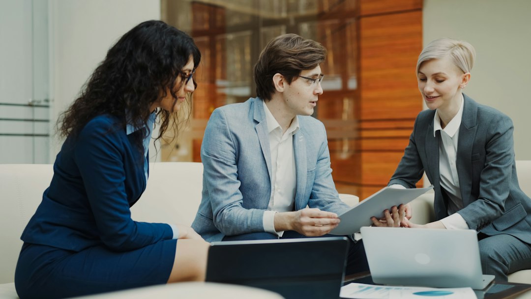 Three coworkers in business attire sit on a sofa, reviewing a document together with laptops nearby.