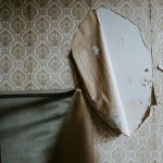 Corner of an abandoned room with ornate beige wallpaper peeling away and a curved, curling sheet of wall material exposing pale blue plaster behind it.