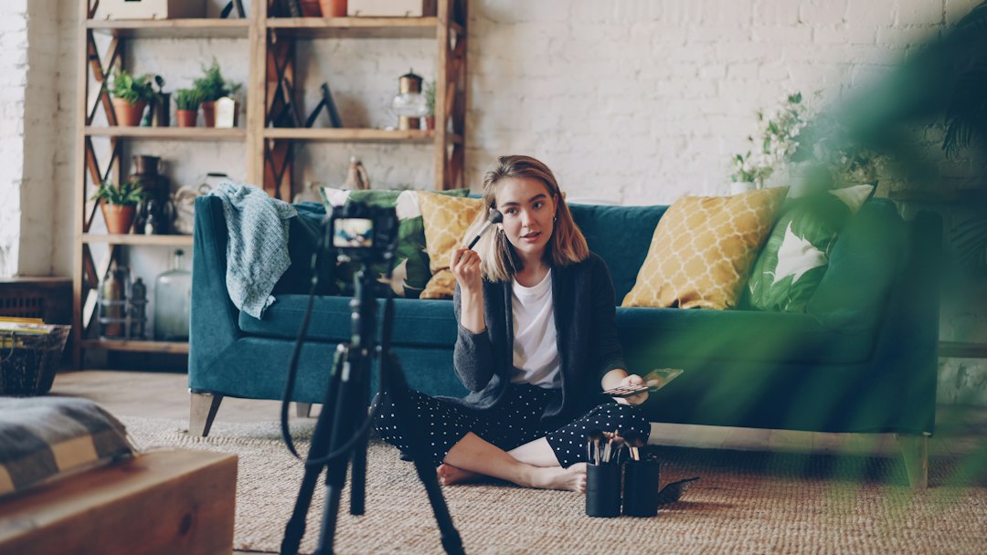 Woman filming a beauty tutorial at home with a camera on a tripod, sitting on the floor beside a blue sofa with plants and cushions.