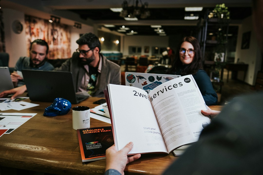 Person holds an open magazine on a wooden table while three coworkers with laptops sit nearby in a modern office.
