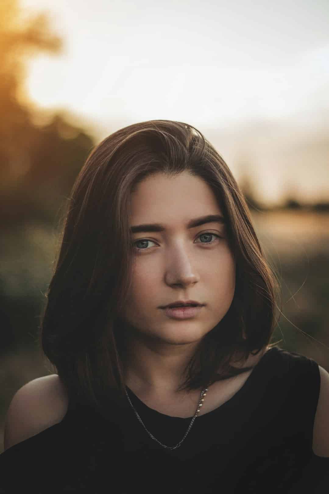 Portrait of a young woman with shoulder-length brown hair and blue eyes, wearing a black top, outdoors at sunset.