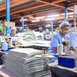 Workers in blue uniforms and hairnets operate sewing machines in a textile factory with tall stacks of finished fabric sheets nearby.