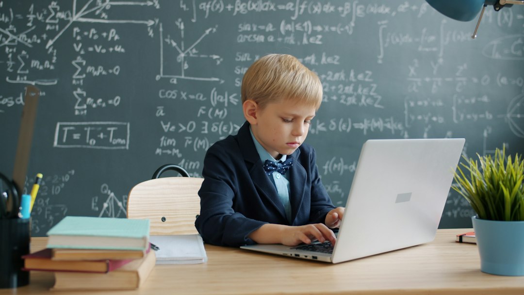 Young boy in a suit typing on a silver laptop at a classroom desk with a chalkboard full of equations behind him.