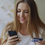 Woman smiling while using a smartphone and holding a credit card.